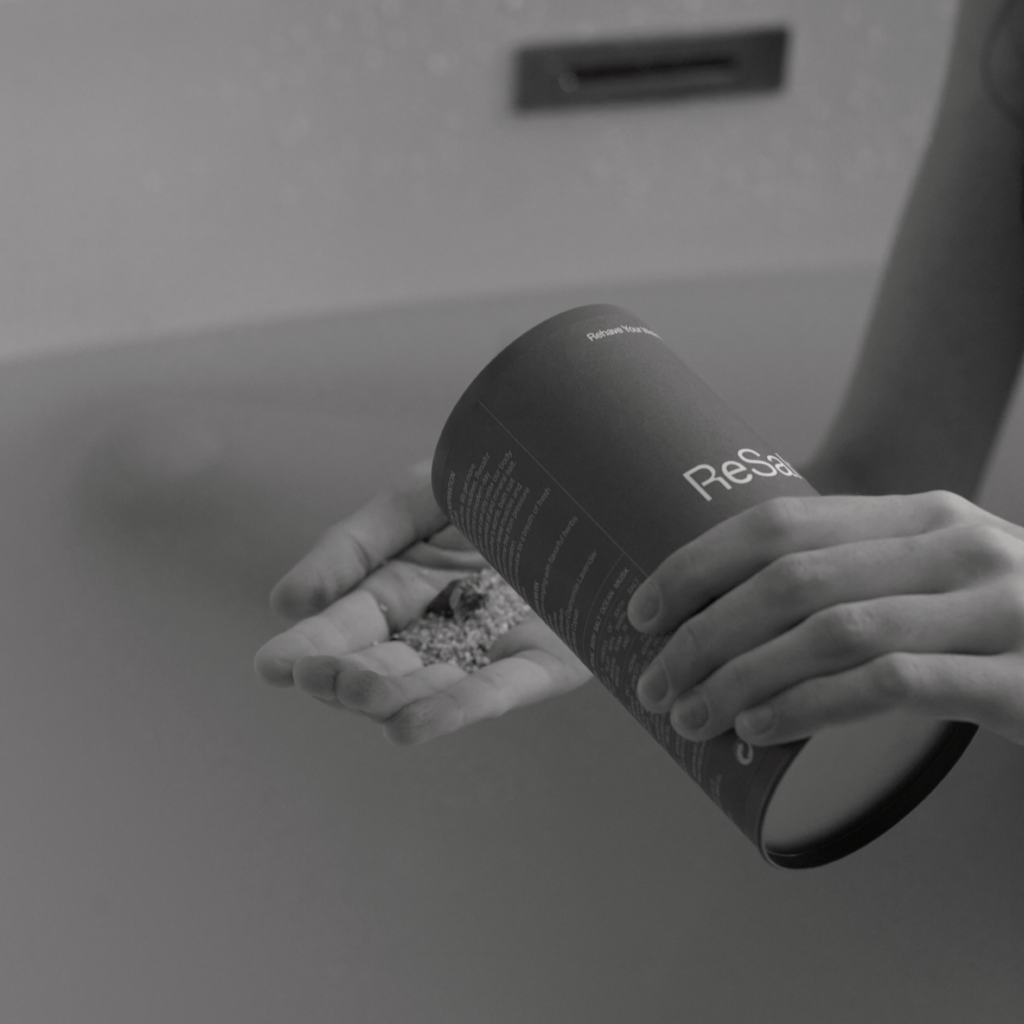 Hands pouring ReSaltZ bath salts from a tin case into a palm, with a bathtub in the background, evoking a calm and relaxing bathing experience.